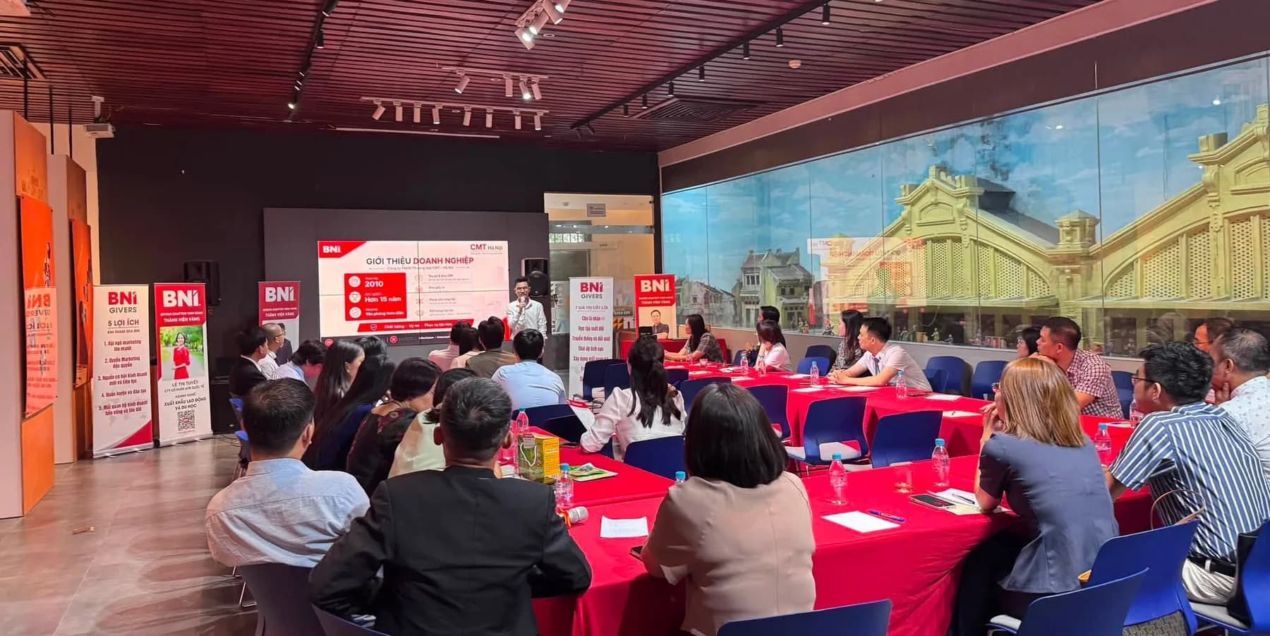 Một thành viên đang trình bày trước phòng họp chapter, các thành viên khác ngồi quanh dãy bàn phủ khăn đỏ